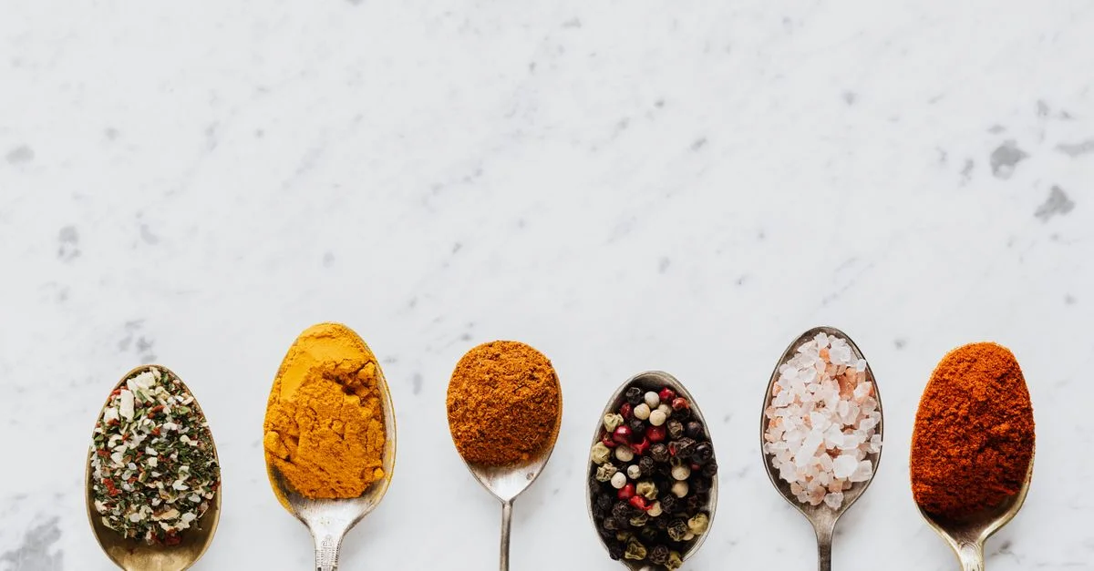 Une variété d'épices et de condiments colorés sur six cuillères disposées sur une surface en marbre.